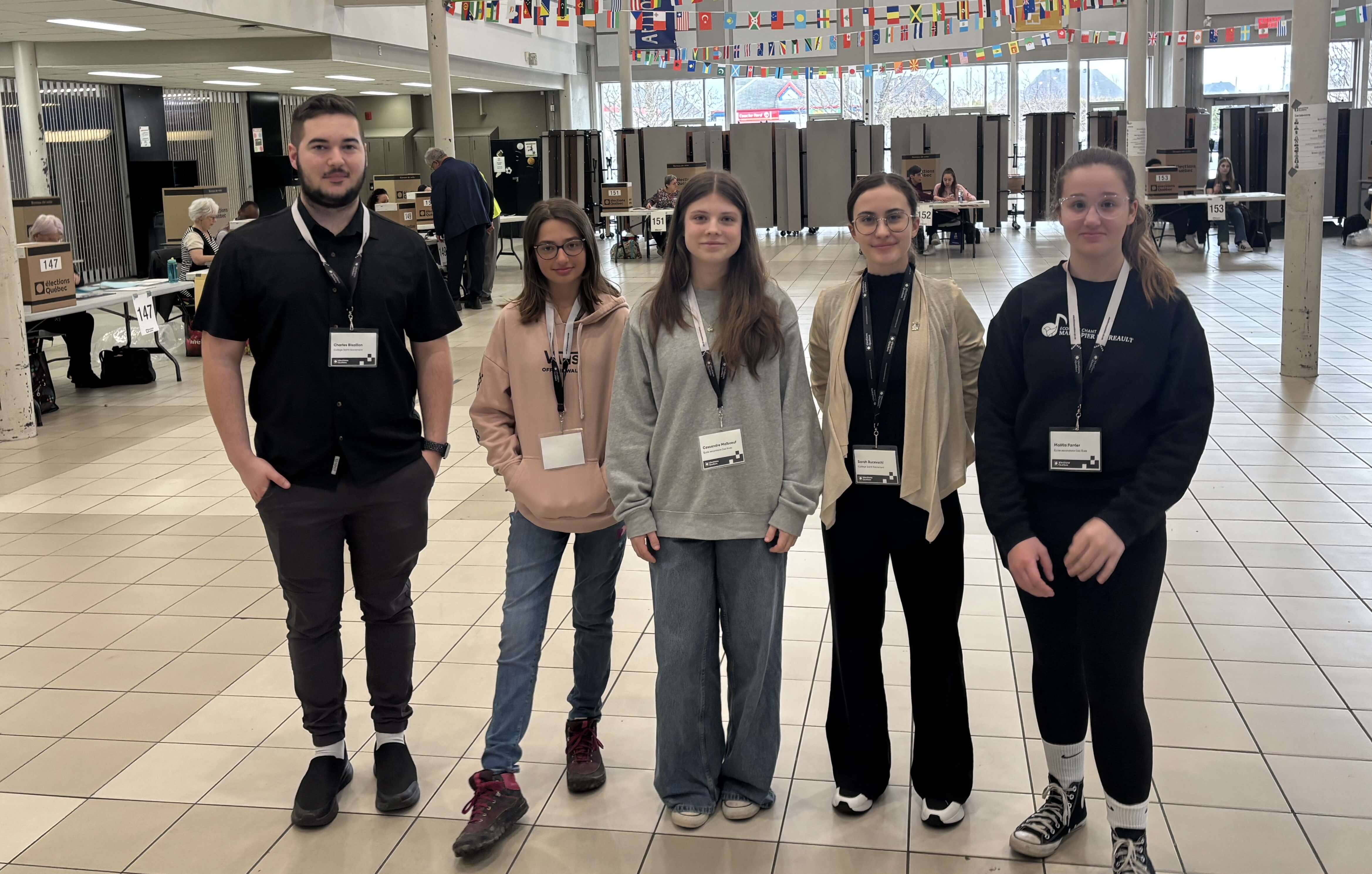 Cinq jeunes dans un bureau de vote entourés du personnel électoral, à leur table respective.
