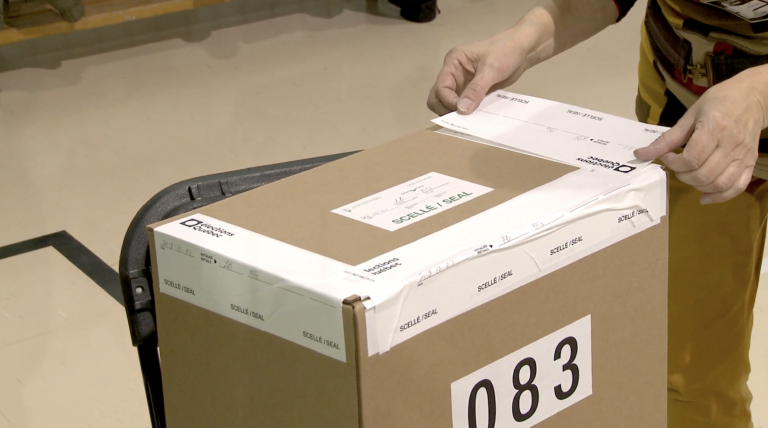 Plan serré d’une urne numérotée avec des autocollants officiels pour la sceller. On voit les mains d’une femme mettant le dernier autocollant nécessaire pour sceller l’urne.